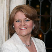 A woman with short light brown hair is smiling, wearing a white jacket and pearl earrings. She stands indoors with a window and dark background behind her, exuding the confidence of a seasoned coach after an invigorating training session.