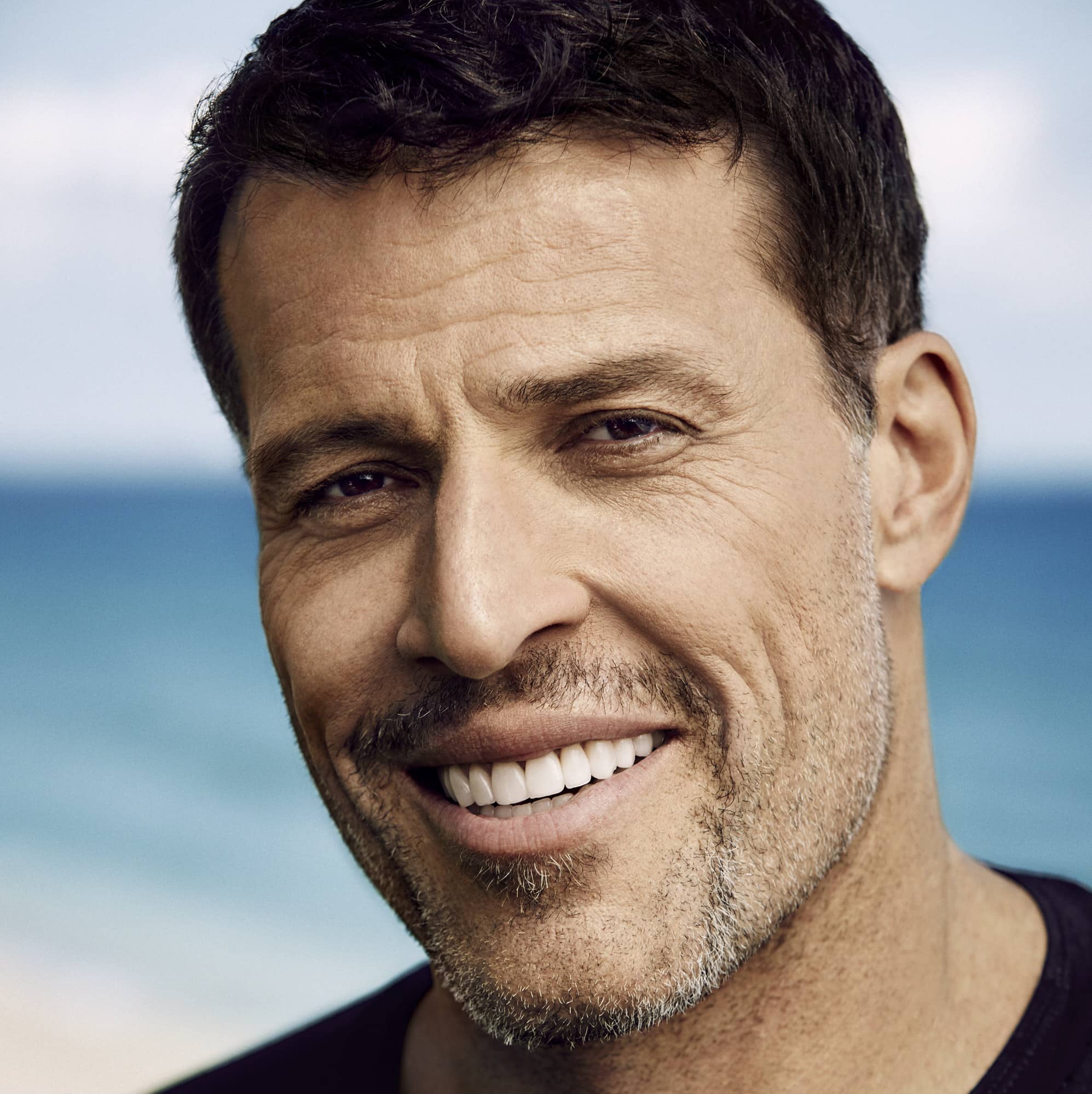 A man named Tony Robbins with short dark hair and slight facial hair smiles, with a beach and ocean visible in the background.