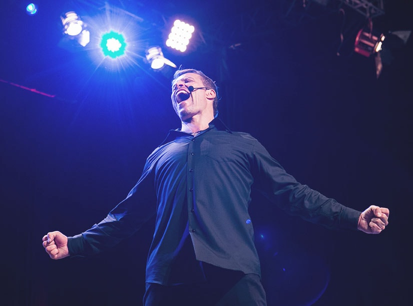 A person in a dark shirt, wearing a headset microphone, raises both arms in a triumphant pose under bright stage lights, showcasing their strategic training.