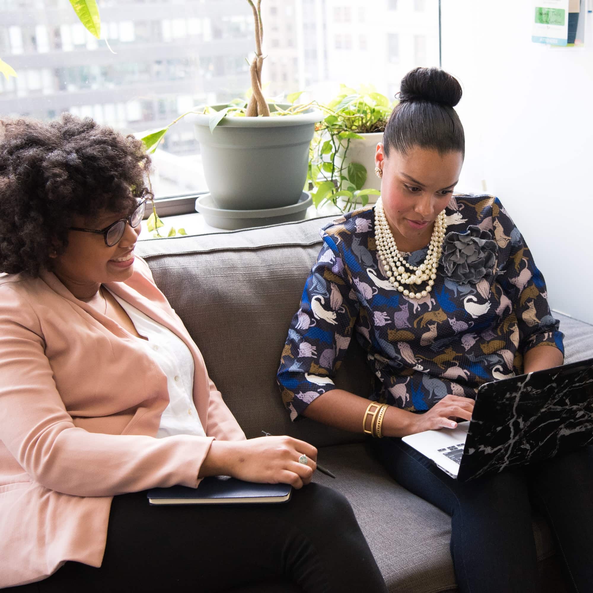 Two women sit on a couch in an office, one with a notebook and the other using a laptop. Plants and a city view are visible in the background, creating a serene setting as they engage in strategic discussions.