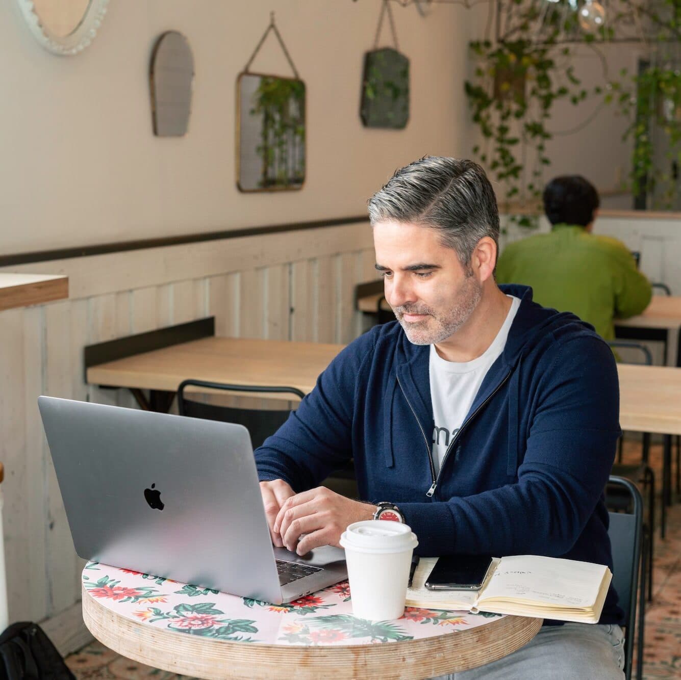 A man with a blue sweater and salt and pepper hair types on a laptop at a small, round table in a cafe. There is a coffee cup and an open notebook beside him. Another person wearing a green shirt is seated in the background.