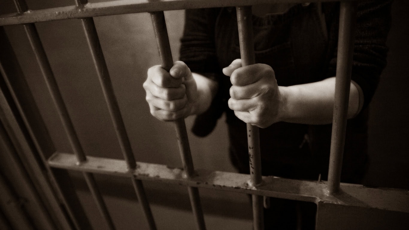 A person is confined to a dimly lit cell, gripping the metal bars with both hands.